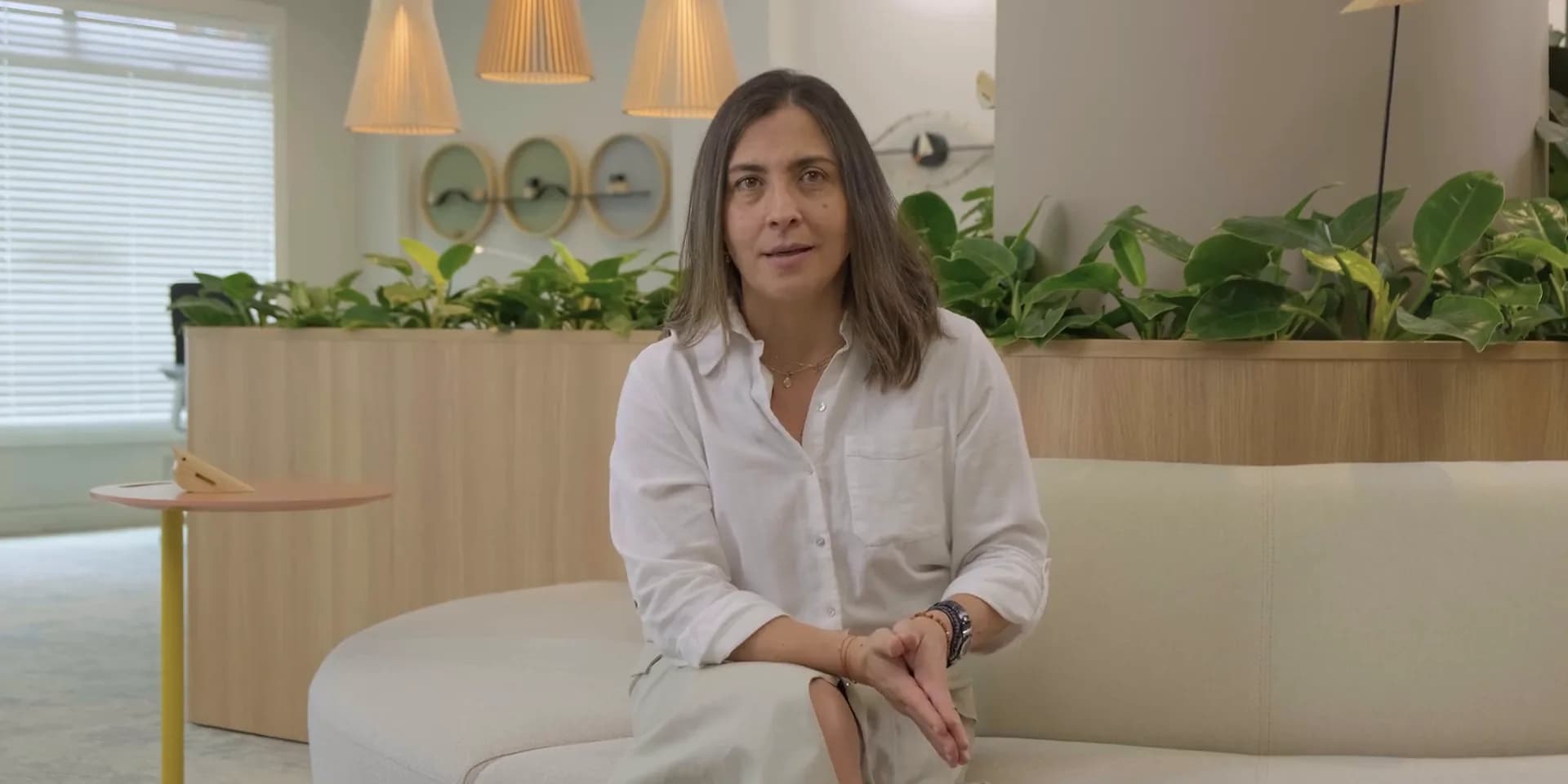 Juliana Arbeláez, sentada y mirando a cámara, en una sala luminosa con plantas y lámparas de mimbre